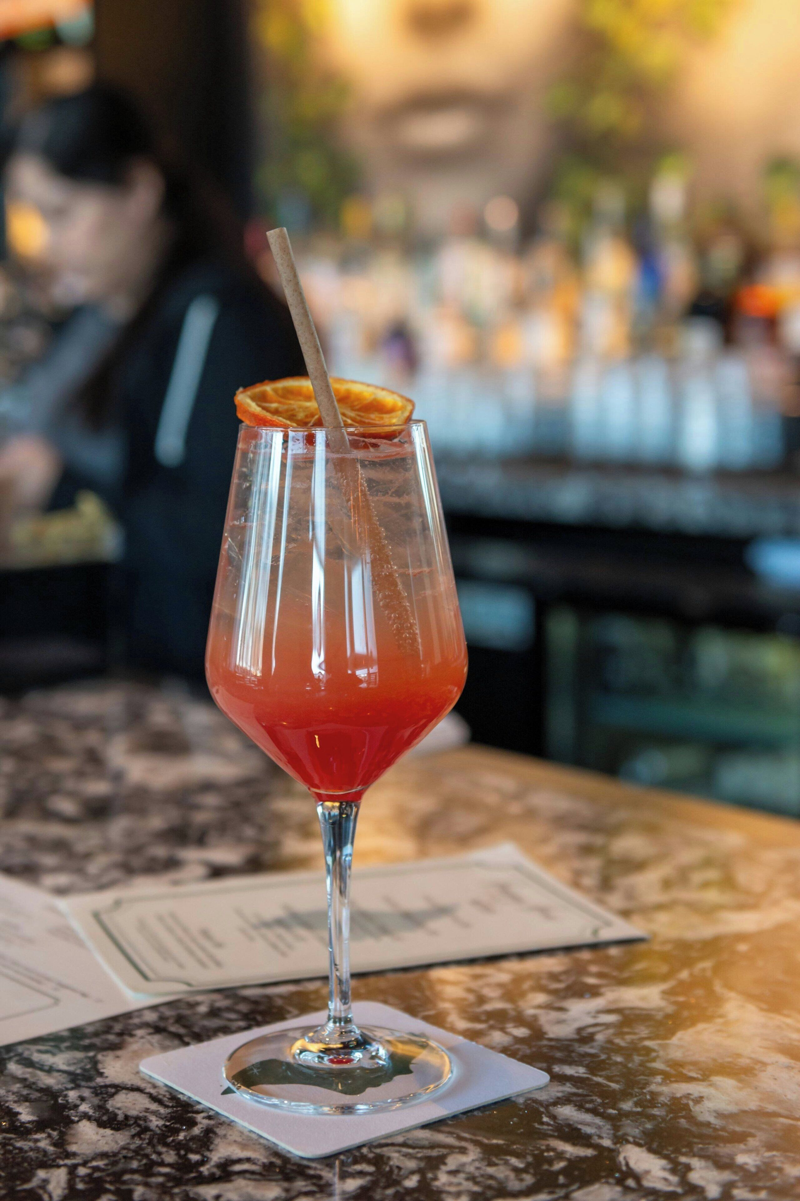 Un vaso de cóctel con pajita y una decoración de gajo de naranja sobre una mesa de mármol, con un fondo desenfocado de una zona de bar.
