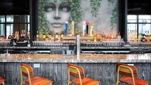Zona de bar con un mural grande, taburetes y una variedad de botellas y vasos sobre la barra.