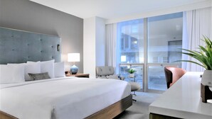 Una habitación de hotel moderna con una cama grande, un escritorio, una silla, una lámpara y vistas a la ciudad a través de amplios ventanales.