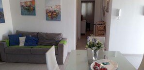 Un salon avec un canapé gris, une table basse en verre et un vase rempli de fleurs. Il y a un tableau au mur et une prise électrique blanche.
