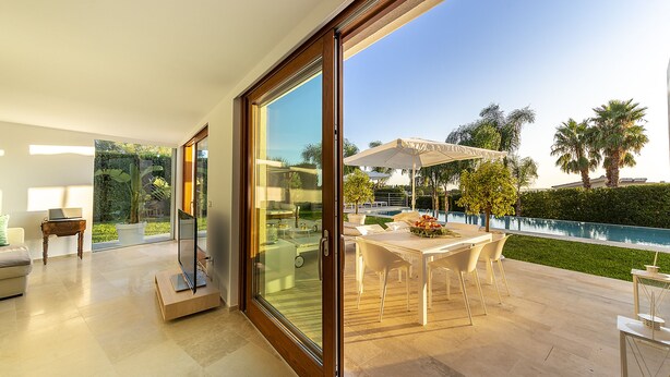 A modern living room with a large sliding glass door leading to an outdoor patio with a pool and palm trees. - Attaláya with swimming pool and garden (San Corrado di Fuori)