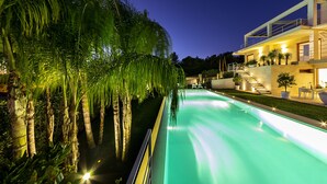 A swimming pool with a water feature, surrounded by palm trees and a modern building with a balcony. - Attaláya with swimming pool and garden (San Corrado di Fuori)
