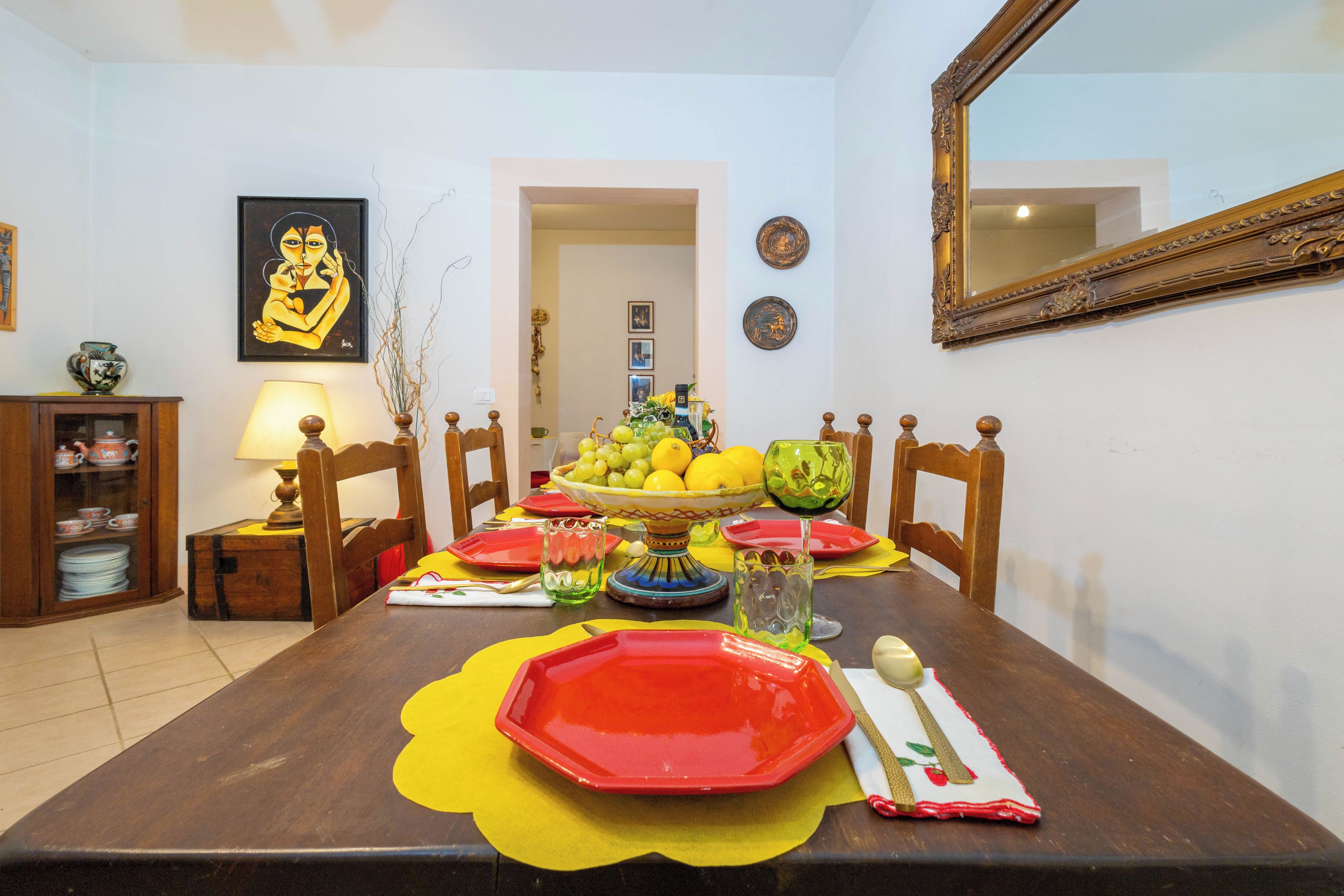 Una mesa de comedor dispuesta con platos rojos, un bol de fruta y un jarrón con flores.