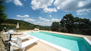 Ein rechteckiger Swimmingpool mit klarem, blauem Wasser, umgeben von einer gefliesten Liegefläche. Weiße Liegestühle und ein einzelner Sonnenschirm sind vorhanden. Im Hintergrund erstreckt sich eine hügelige Landschaft mit Bäumen und blauem Himmel.