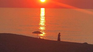 Un coucher de soleil sur une mer calme, avec une personne debout sur la plage, à l’ombre d’un parasol.