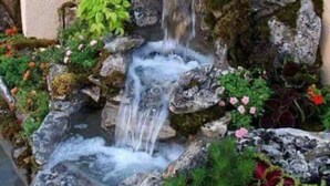 A small waterfall feature with a terracotta pot at the top, surrounded by rocks and various plants. - Villa Corina near the Athens airport (Attica)