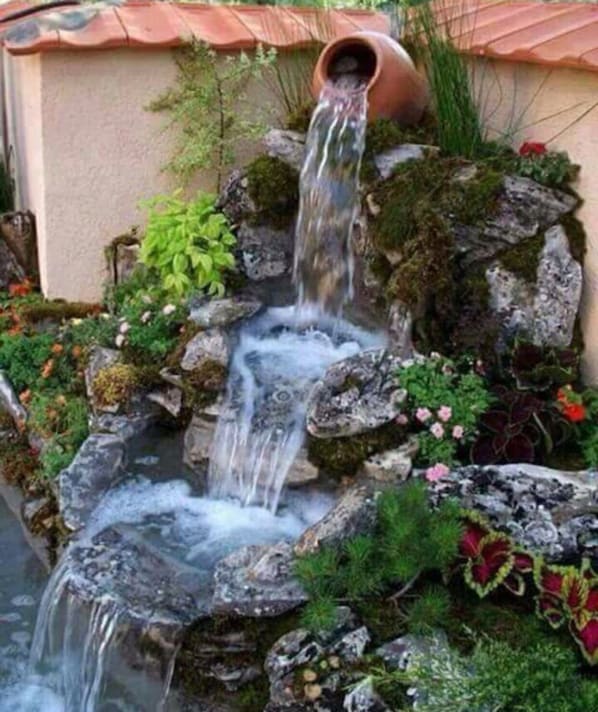 Une petite cascade ornée d’un pot en terre cuite au sommet, entourée de rochers et de différentes plantes.
