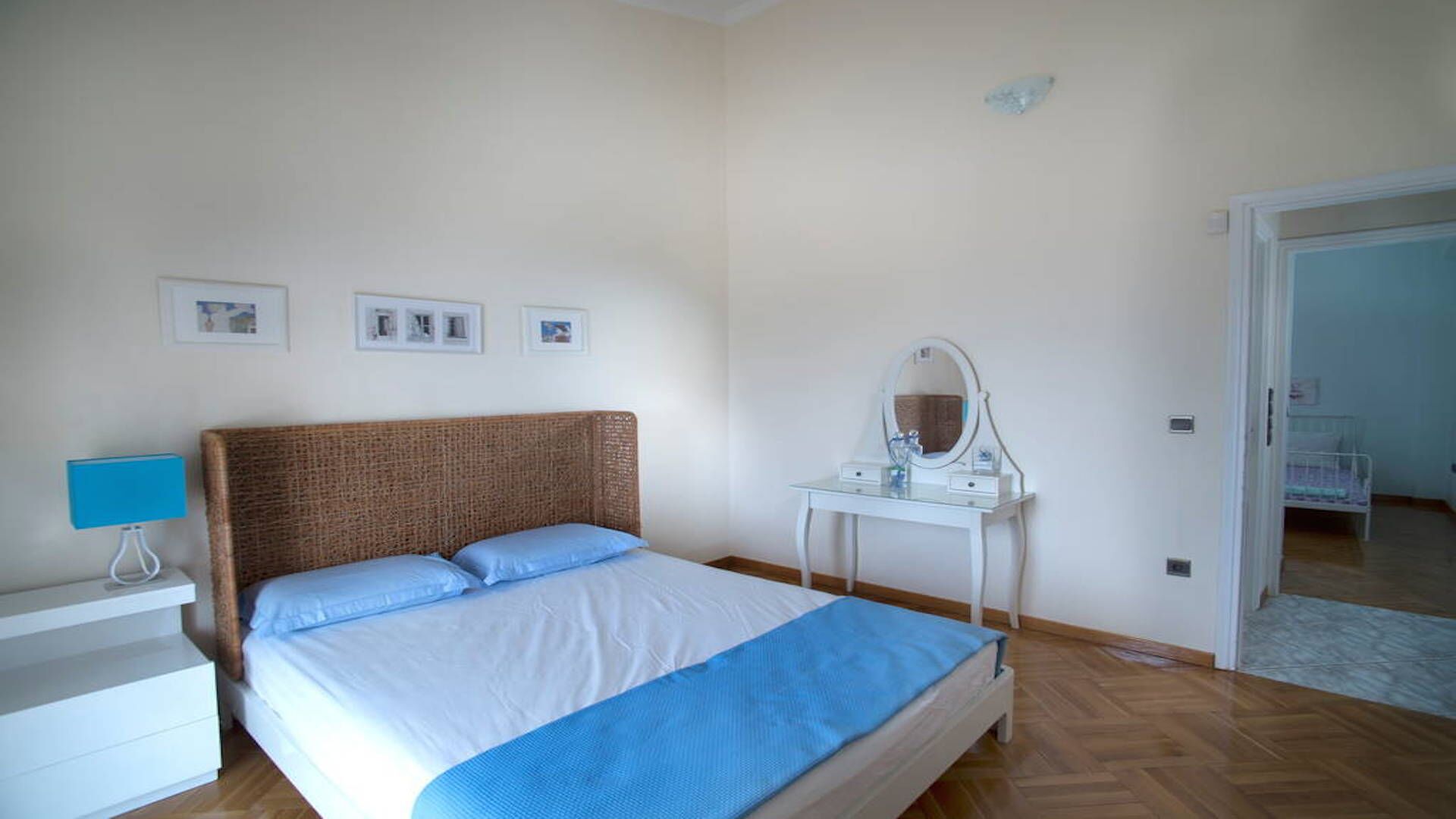A bedroom with a bed, a bedside table with a lamp, a dresser, and a door leading to another room.