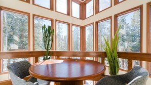 A round wooden table with two grey chairs, surrounded by large windows with wooden frames, overlooking a forested area.