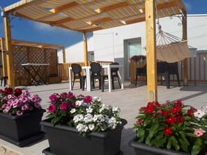 Eine Terrass mit Holz-Pergola, schwarzen Stühlen, einem Tisch und Kübelblumen.