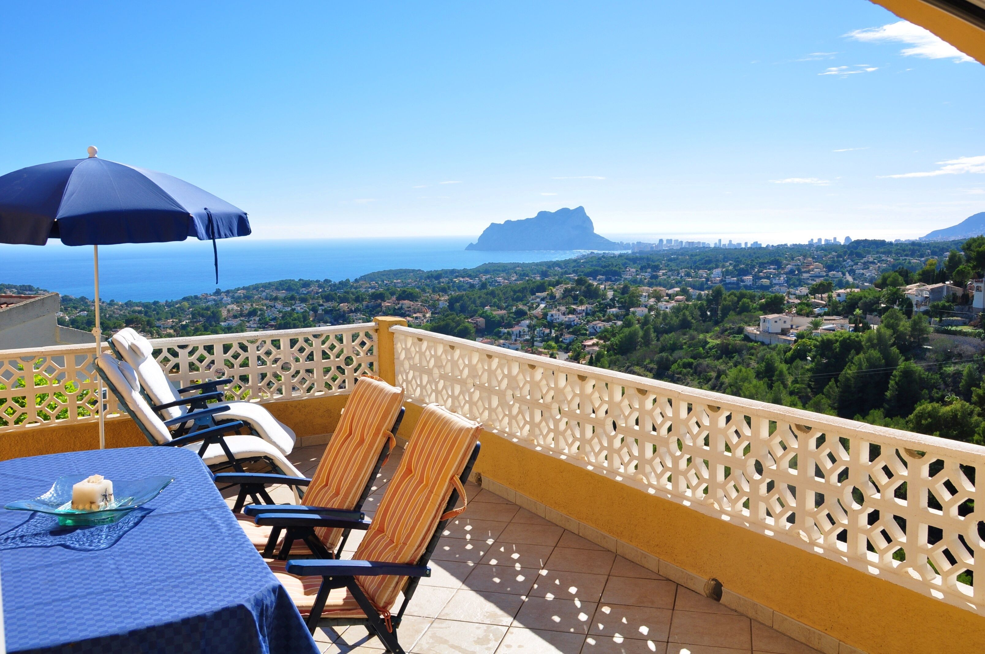 Varanda com mesa, cadeiras e guarda-sol, com vista para uma cidade costeira e o mar.