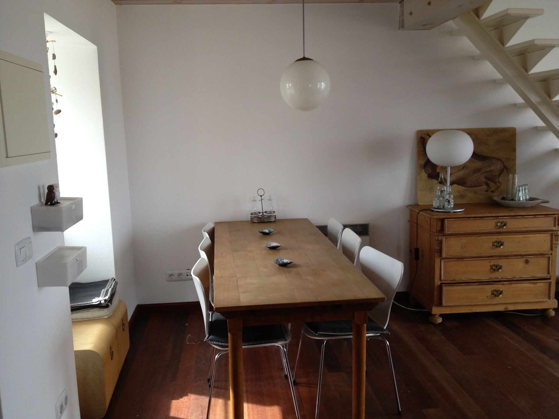 A dining area with a wooden table, white chairs, a hanging light, a wooden dresser, and a painting on the wall.