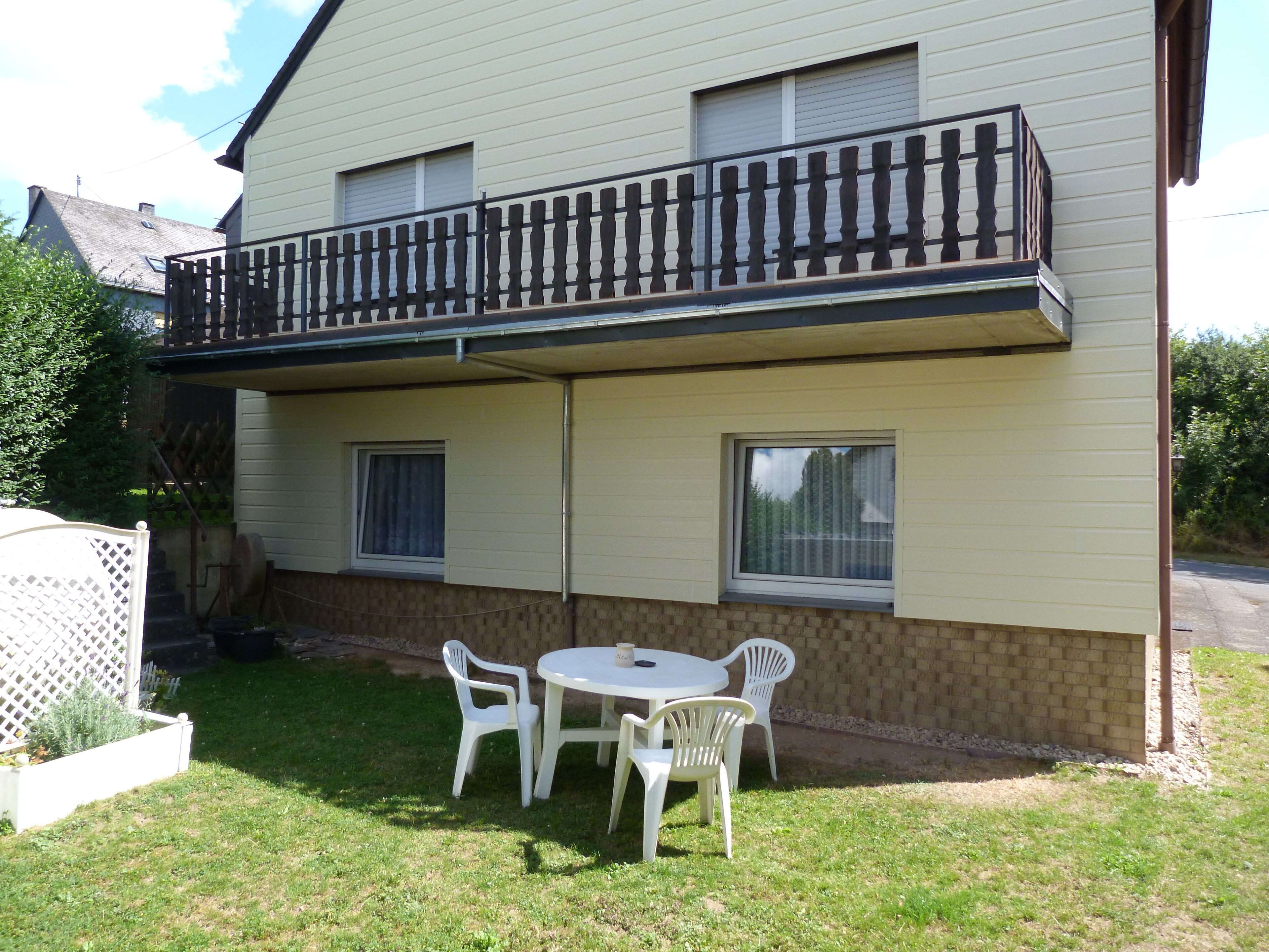 A two-story house with a balcony, a small patio with a table and chairs, and a garden area.