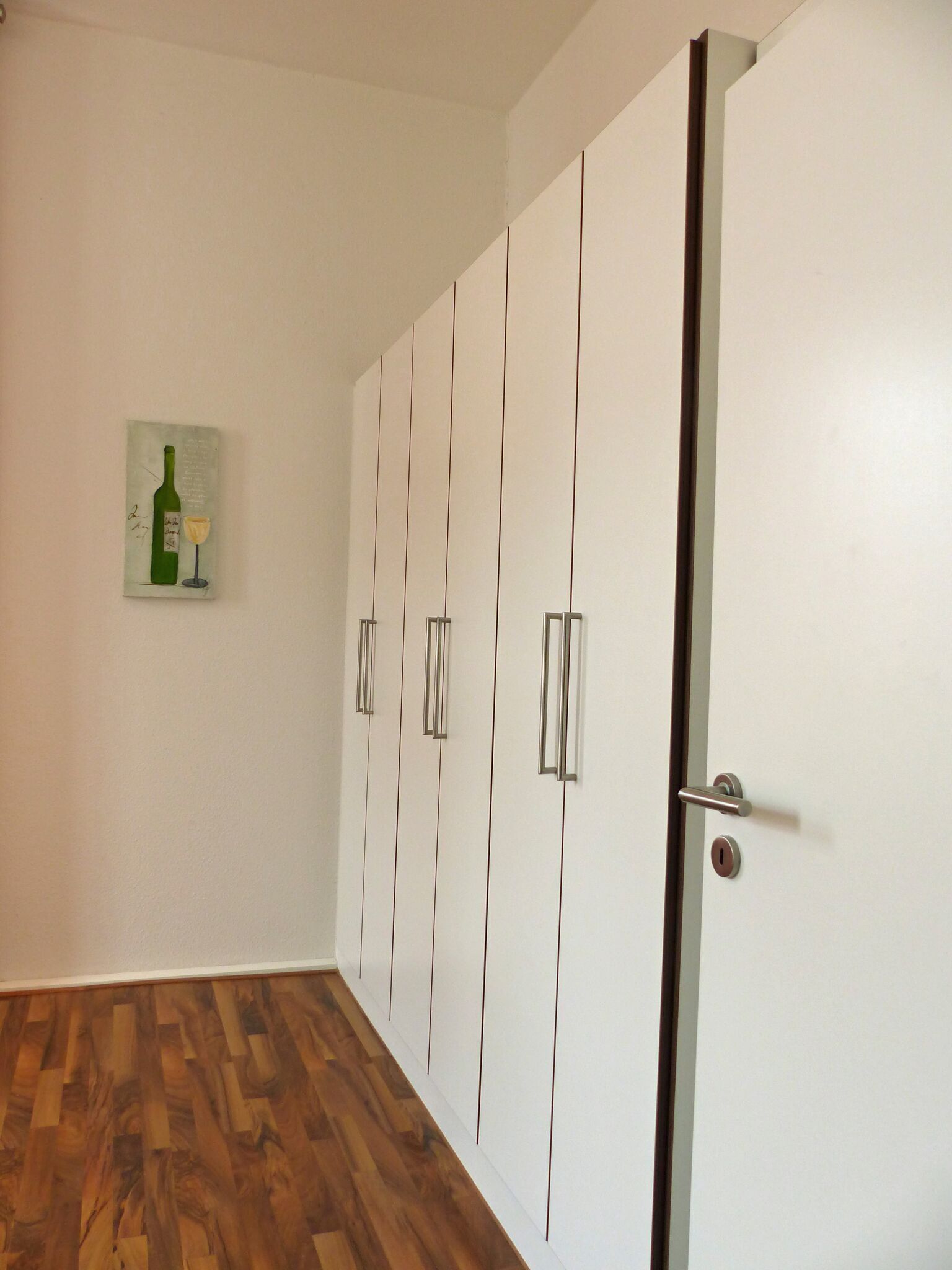 Ein Zimmer mit Holzboden, einer weißen Tür mit silbernem Griff und einem an der Wand hängenden Kunstwerk, das eine Weinflasche und ein Weinglas zeigt.