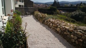 Un camino de grava que conduce a un muro de piedra, con un edificio a la izquierda y vistas de un pueblo y colinas a lo lejos.