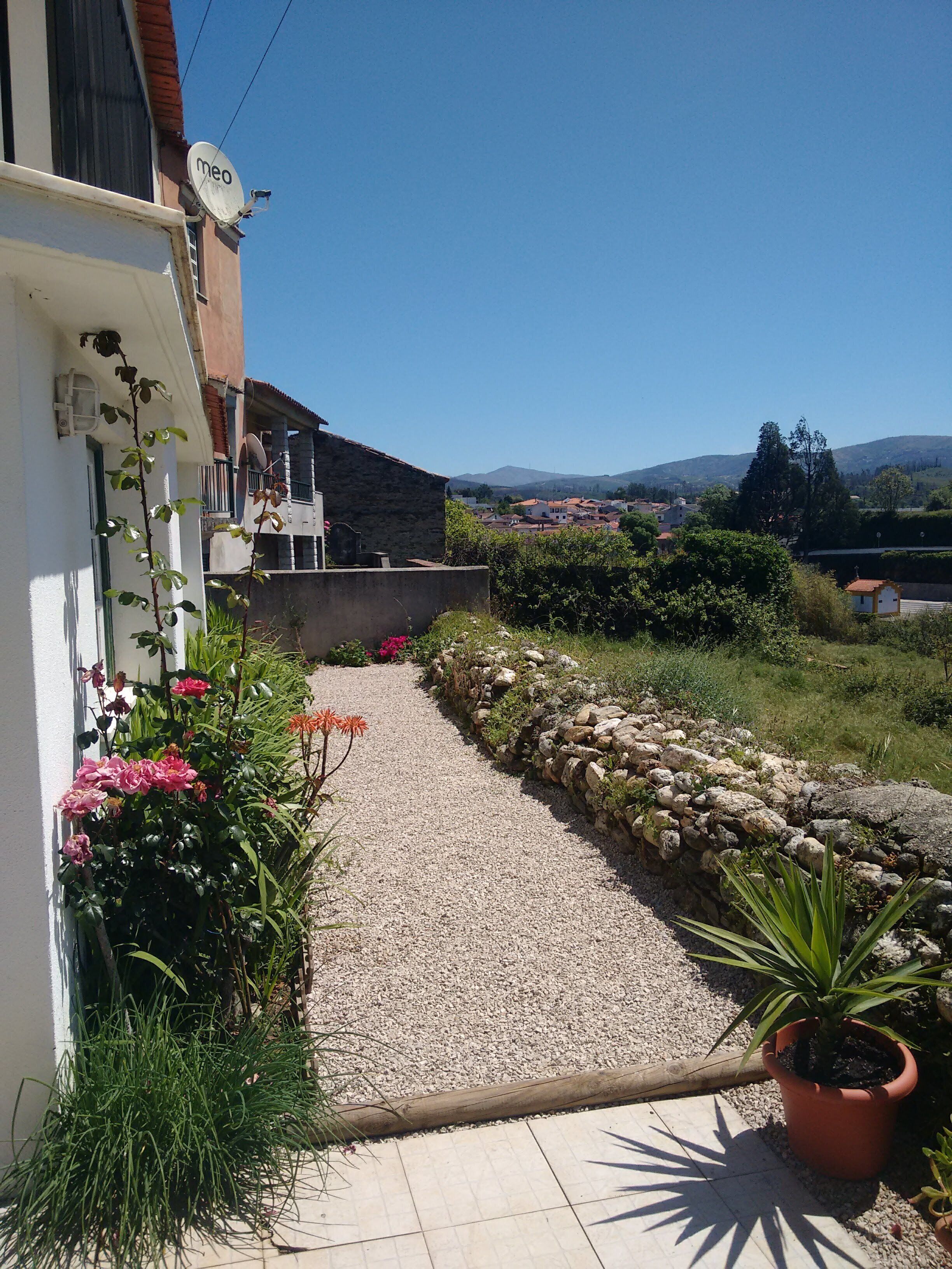 A gravel pathway leading to a building with a satellite dish, surrounded by greenery and a view of distant hills.