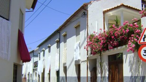 Una calle estrecha flanqueada por edificios blancos, algunos con balcones y flores. Se ve una señal de límite de velocidad.