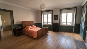Quarto com cama de madeira, mesa de cabeceira, radiador e janelas amplas.