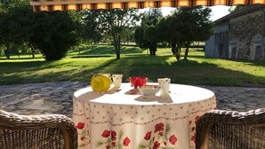 A round table with a floral tablecloth, surrounded by wicker chairs, set outdoors under a striped awning.