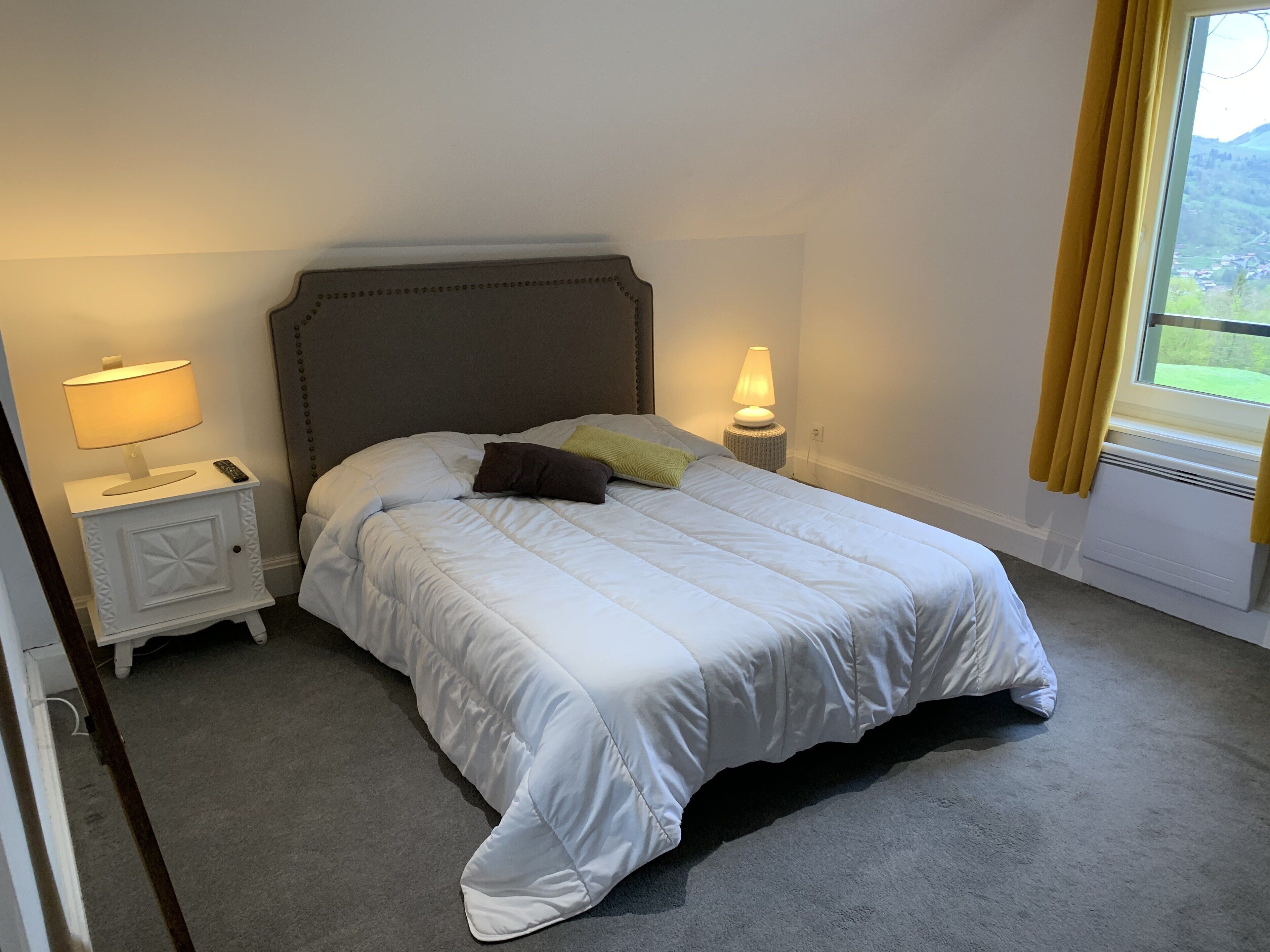 A neatly made bed with white bedding, two bedside lamps, a small bedside table with a lamp, and a window with curtains.