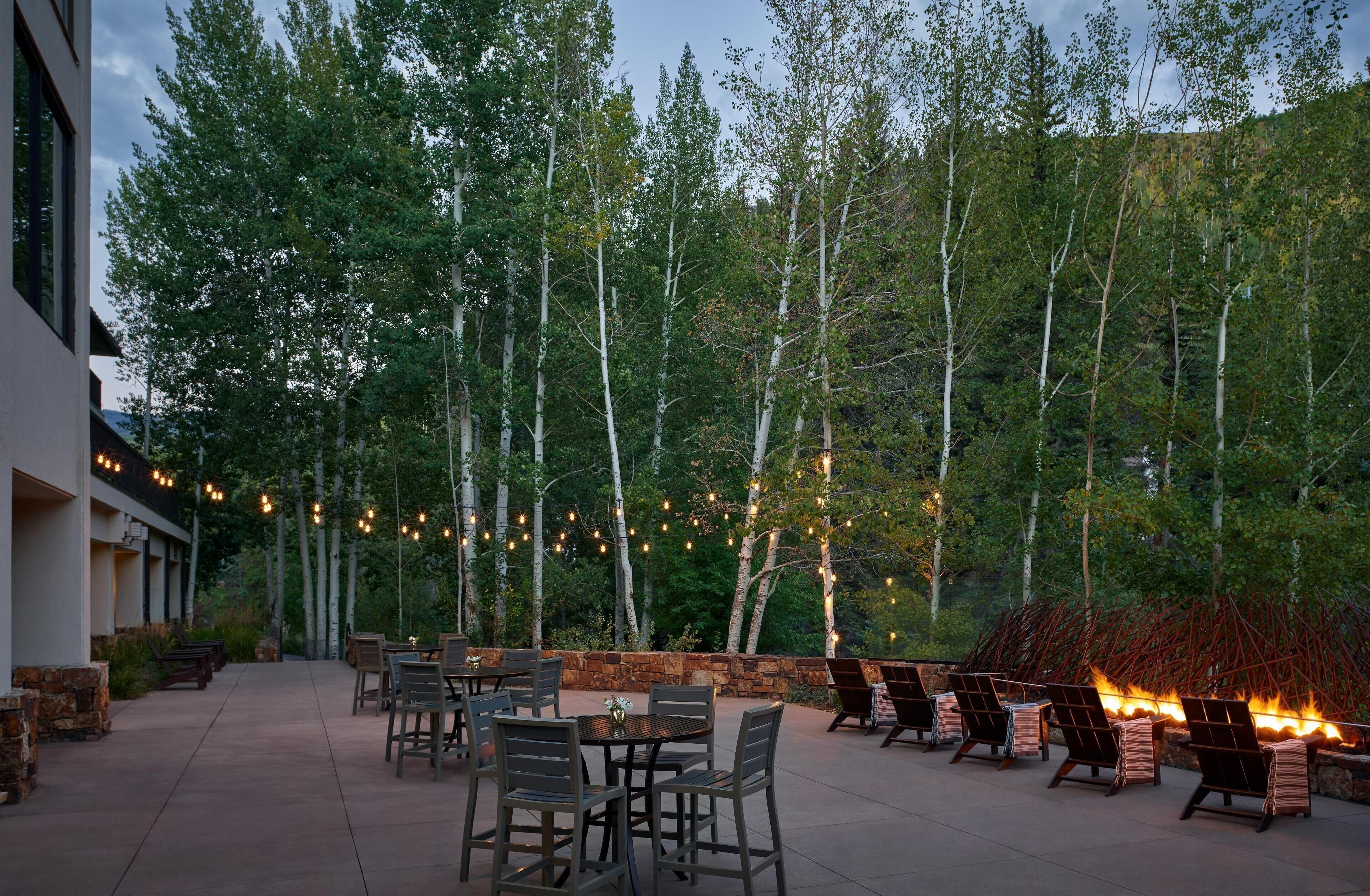 An outdoor patio with tables and chairs, surrounded by trees and a stone wall.