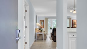 Un pasillo con una puerta blanca, una mesita con lámpara, un sofá y una silla. Se ve una ventana con cortinas y vistas a árboles.