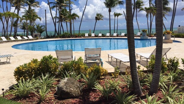 A pool surrounded by lounge chairs and palm trees, with a view of the ocean. - Ocean Front - 3 Bedroom/3.5 Bath with Terrace Fully Furnished (Humacao)