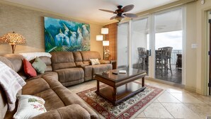 A living room with a brown leather sofa, a glass coffee table, and a painting of birds. There is a sliding glass door leading to an outdoor area with chairs. - Gulf Views! (Pensacola)