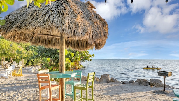 A beach setting with a thatched umbrella, wooden chairs, and a table.