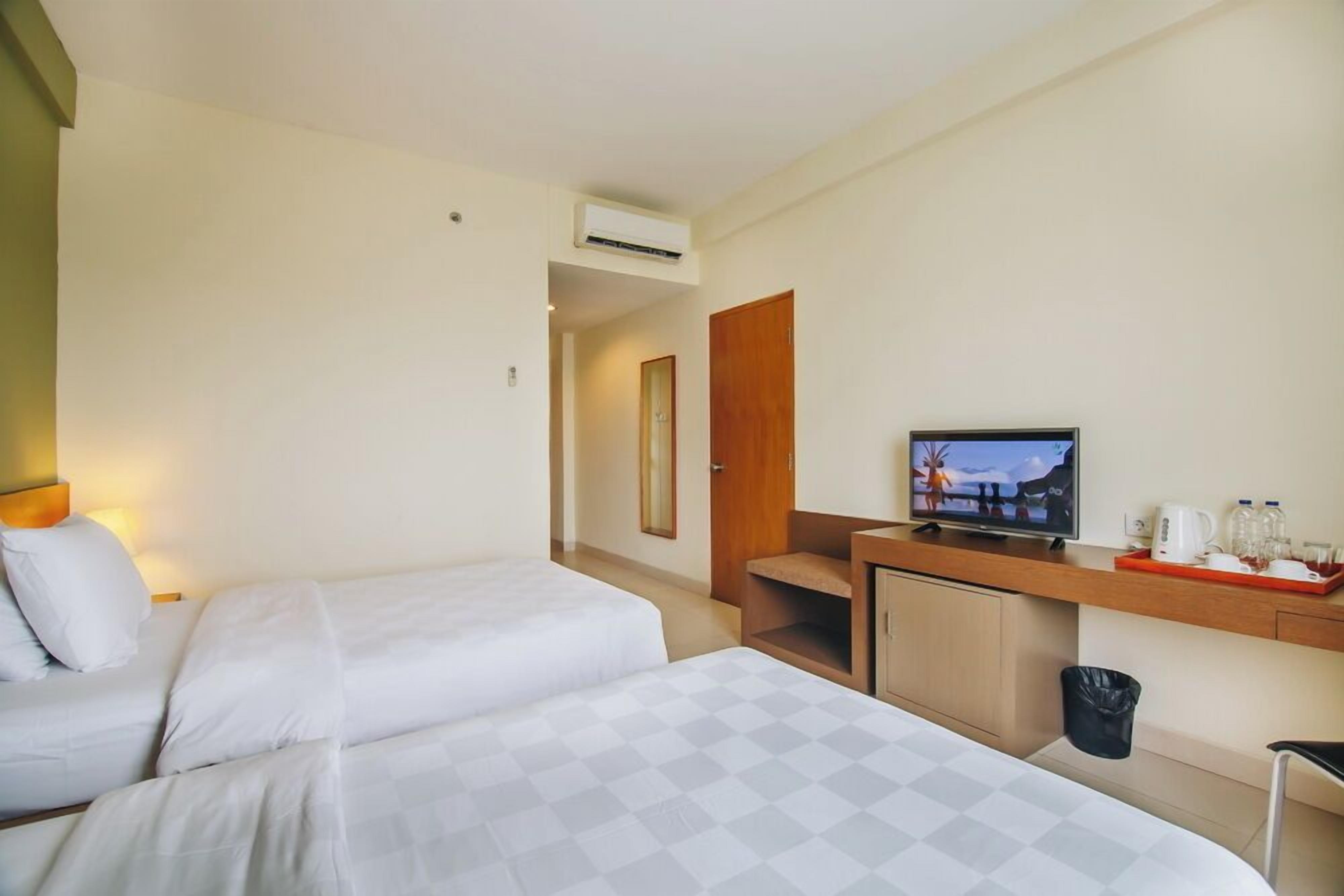 A hotel room with two beds, a desk with a television, a small kitchenette, and a trash bin.