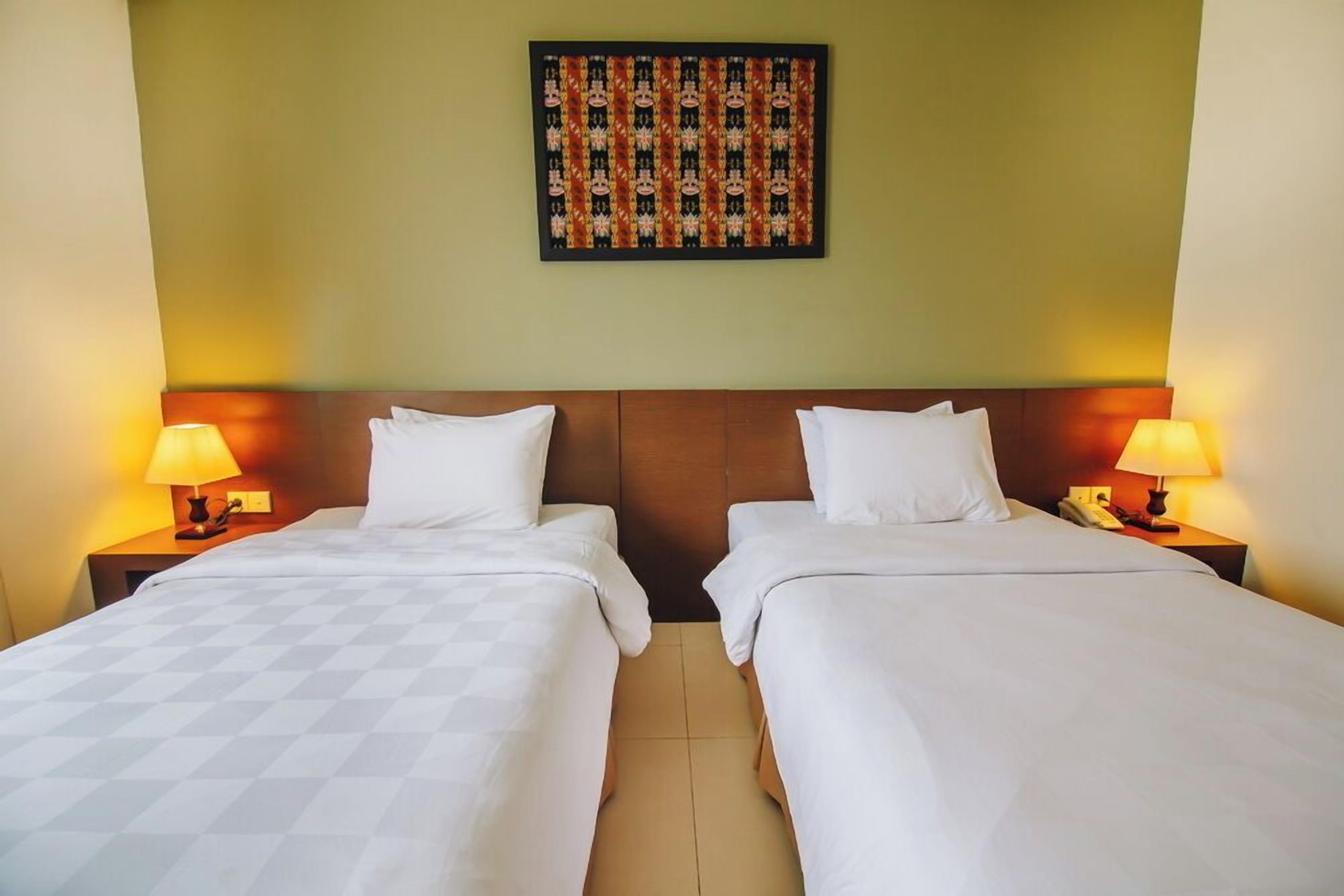 Una habitación de hotel con dos camas, cada una con ropa blanca y cabeceros de madera. Hay lámparas de noche a cada lado de las camas.