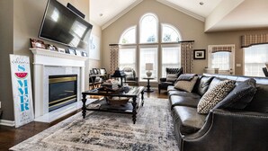 A spacious living room with a fireplace, a large TV mounted above it, and a comfortable seating area with a leather couch and a coffee table. - Beautiful 4 BR, 4.5 Bath, Executive Home w In Ground Pool & Handicap Accessible! (Louisville)