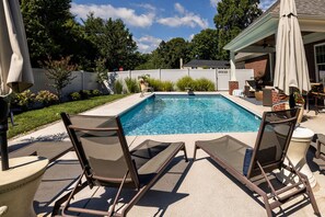 Ein rechteckiger Swimmingpool mit klarem, blauem Wasser, umgeben von einer Betonplattform. Zwei Liegestühle mit Metallgestellen und Netzbespannung sind vorhanden.