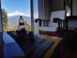 Una habitación con vistas a montañas nevadas, una silla de madera, una mesa con una botella, un vaso y una taza.