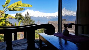 A view through a window with a mountain backdrop, a wooden chair, and a vase with flowers on a table. - ES Deurali Resort. (Pokhara)