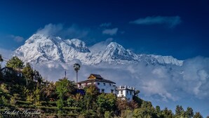 A white building with a traditional architectural style, surrounded by lush greenery and a backdrop of snow-capped mountains. - ES Deurali Resort. (Pokhara)
