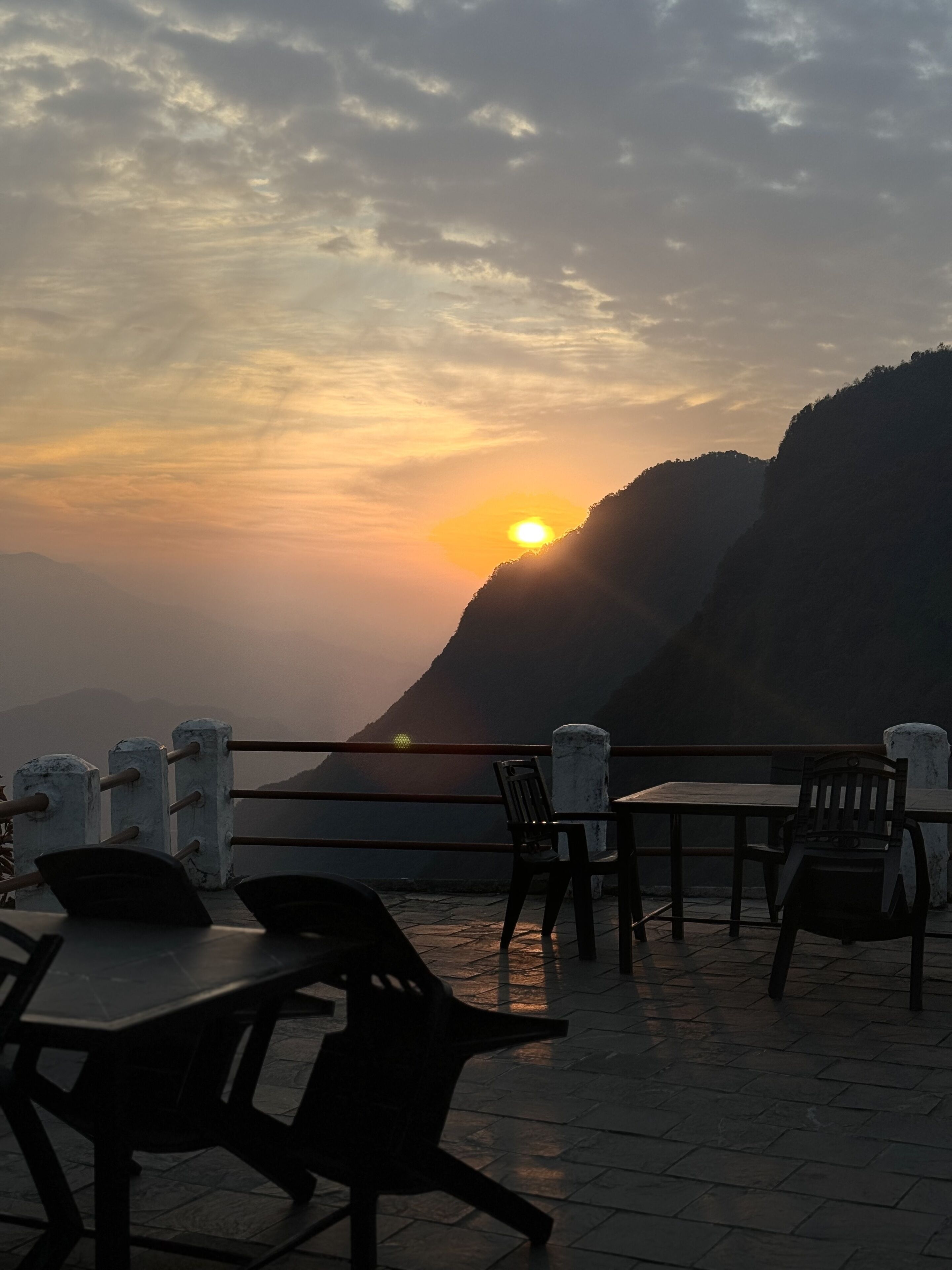 A terrace with tables and chairs, overlooking a mountainous landscape during sunset.