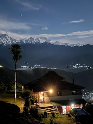 A mountain lodge with a view of snow-capped peaks, a palm tree, and a lit entrance. - ES Deurali Resort. (Pokhara)
