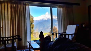 A room with a large window offering a mountain view. There is a bed with a person lying down, a chair, and a table. - ES Deurali Resort. (Pokhara)