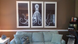 A living room with a light blue sofa, a framed octopus artwork on the wall, and a bookshelf with books and a globe. - Gated Community: Top Corner Unit, Screened Porch Facing Woods, Beautiful Pool (Rehoboth Beach)