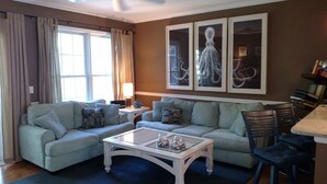 A living room with a light blue sofa set, a glass coffee table, and a dining area with a table and chairs. - Gated Community: Top Corner Unit, Screened Porch Facing Woods, Beautiful Pool (Rehoboth Beach)