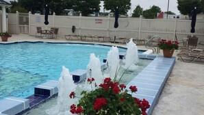 A swimming pool with water fountains, potted red flowers, and a white fence. - Gated Community: Top Corner Unit, Screened Porch Facing Woods, Beautiful Pool (Rehoboth Beach)