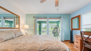 A bedroom with a large bed, a desk, and a chair. There is a window with curtains and a view of greenery outside.