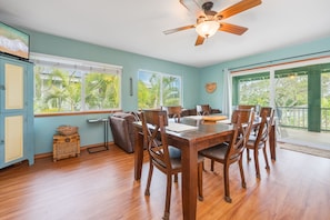 Un coin repas avec une table et des chaises en bois, un ventilateur de plafond et de grandes fenêtres donnant sur des palmiers.