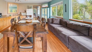 A spacious living area with a dining table, wooden chairs, a gray sofa, and large windows offering a view of palm trees.