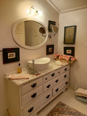 A bathroom with a white sink, a large mirror, and a vanity with multiple drawers. There are framed pictures on the wall and a small vase with flowers on the vanity.