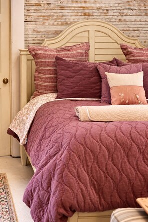 Un lit avec une couette violette matelassée et plusieurs oreillers, dont un orné d’un motif floral.