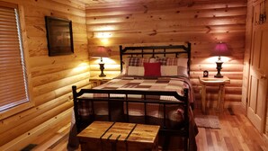 A cozy wooden cabin bedroom with a bed, bedside lamps, a nightstand, a wooden chest, and a window with blinds. - Beautiful cabin style home backing up on the White River! (Mountain View)