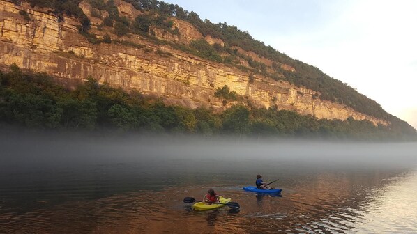 Un río tranquilo con kayaks y una persona remando, rodeado de acantilados y niebla.