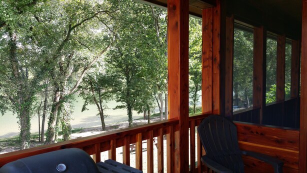 A wooden deck with a grill and a chair, overlooking a lake surrounded by trees. - Beautiful cabin style home backing up on the White River! (Mountain View)